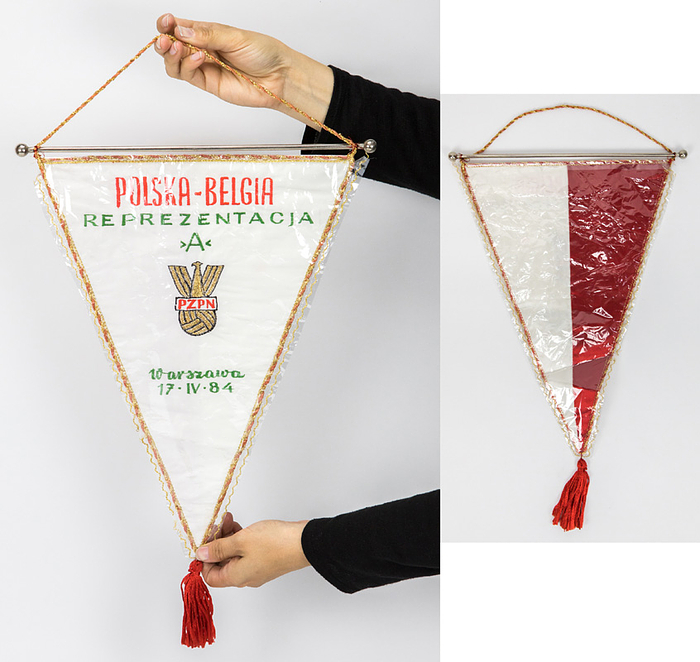  - Official cotton match pennant of the Polish national team, featuring the embroidered inscription "Polska - Belgia Reprezentacja A. Warszawa 17 IV 1984" (Poland vs. Belgium friendly match, 0–1). The front is partially embroidered with gold brocade; the back displays the Polish national colors. The side borders feature gold brocade and silk. Includes a silk tassel and a chrome rod with ball finials; dimensions: 38 x 28 cm (excluding tassel). The pennant comes in its original cellophane sleeve, which is stitched with gold brocade thread. This pennant was presented to the Belgian team captain, Jean-Marie Pfaff, in the center circle! --The pennant comes directly from Jean-Marie Pfaff's collection. Pfaff's collection was auctioned off in Paris on May 23, 2026! The consignor purchased this pennant at that auction. Condition: A-