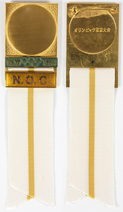 Teilnehmerabzeichen 1964  | Offizielles Teilnehmerabzeichen „N.O.C.“ (Nationales Olympisches Komitee) für die Olympischen Spiele Tokyo 1964 .  Bronze, vergoldet. Schriftleiste golden emailliert. Seidenband in weiß-gelb. 15x3,8cm.