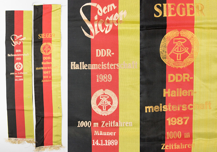 Meisterscherpen DDR 1987 + 1989  | Zwei Meisterscherpen der DDR Bahnradsport Meisterschaften: 1) "Sieger DDR - Hallenmeisterschaft 1987 1000 m Zeitfahren" (Gewinner Lutz Heßlich 2x Gold bei den Olympischen Spielen 1980 + 1988; 4x Weltmeister). Mit kleiner Beschädgung. 2) "Dem Sieger DDR - Hallenmeisterschaft 1989 1000 m Zeitfahren Männer 14.1.1989" (Michael Hübner, 7facher Weltmeister). Beide Kreppstoff mit goldbedruckter Aufschrift. Seidenfransen. 104x19,5 + 92x38 cm. Beides Unikate!