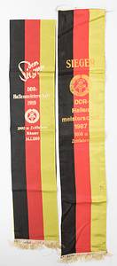 Meisterscherpen DDR 1987 + 1989  | Zwei Meisterscherpen der DDR Bahnradsport Meisterschaften: 1) "Sieger DDR - Hallenmeisterschaft 1987 1000 m Zeitfahren" (Gewinner Lutz Heßlich 2x Gold bei den Olympischen Spielen 1980 + 1988; 4x Weltmeister). Mit kleiner Beschädgung. 2) "Dem Sieger DDR - Hallenmeisterschaft 1989 1000 m Zeitfahren Männer 14.1.1989" (Michael Hübner, 7facher Weltmeister). Beide Kreppstoff mit goldbedruckter Aufschrift. Seidenfransen. 104x19,5 + 92x38 cm. Beides Unikate! - Bild 2
