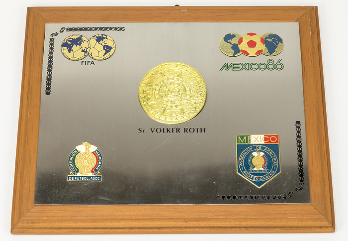 Ehrenplakette WM 1986  | Große Ehrenplakette des mexikanischen Fußballverbandes und der mexikanischen Schiedsrichtervereinigung für die Schiedsrichter der Fußball - Weltmeisterschaft 1986. Persönliches Exemplar von "Volker Roth" (mit Namensgraur). Mit vier aufgesetzten farbig emaillierten Plaketten der FIFA, Logo "Mexico 86", des mexikanischen Fußballverbandes und des mexikanischen Schiedsrichterverbands auf einer Edelstahlplatte, die auf einer Holzplatte montiert wurde, 33x28 cm