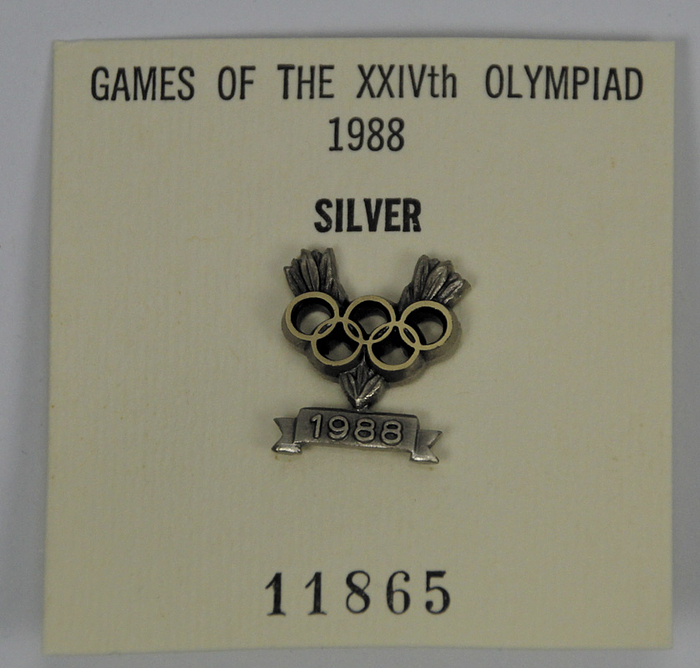 Siegerpin-IOC 1988  | Ehrennadel des I.O.C. für Silbermedaillengewinner bei den Olympischen Sommerspielen Seoul 1988. Persönliches nummeriertes Abzeichen des deutschen Fechters Ulrich Schreck für den 2.Platz mit der Mannschaft. Nummer "11865". Bronze, versilbert. 2x1,4 cm. In Originaletui.