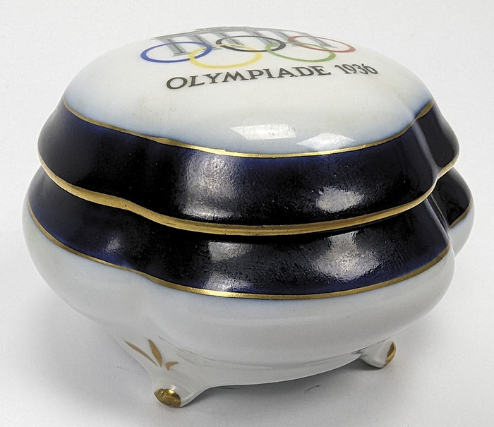 Olympic Games 1936. Commemorative Porcelain Box - Porcelain lidded box with the Brandenburg Gate, colored Olympic rings and the inscription "Olympics 1936". Manufacturer: "PM". Diameter: 8.2cm; Height. 6cm --Very rare, decorative souvenir piece for the 1936 Olympic Games. Inside with one small chip -- Condition: A-