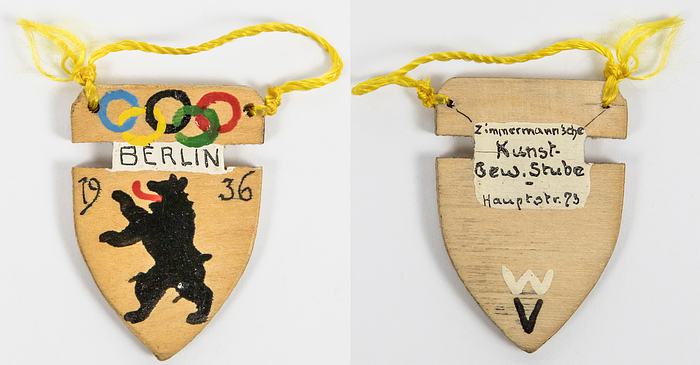 Handgefertiges Souvenirabzeichen für die Olympischen Spiele Berlin 1936. Handbemaltes Holzabzeichen mit den Olympischen Ringen, den Berliner Bär und der Aufschrift "Verlin 1936". 3,5x3,5 cm mit original Seidenbändchen. Hersteller: Zimmermansche Kunst Gewerbe-Stube Hauptstr. 23".