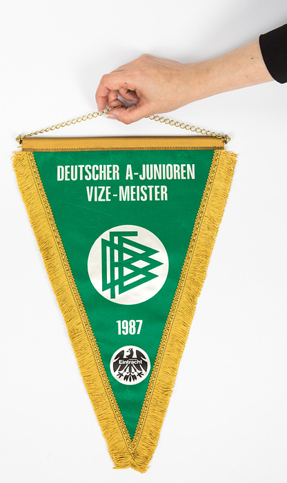 Offizieller Meisterschaftswimpel des DFB für die A-Jugend von Eintracht Frankfurt 1987 mit aufgedrucktem dem Logo des DFB und von Eintracht Frankfurt und der Aufschrift "Deutscher A-Junioren Vize-Meister 1987". Stoff mit Seidenfransen, Messingstange mit Kette, 42x30 cm.