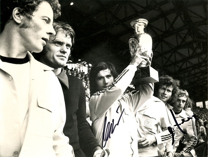 (1945-2021) s/w-Pressefoto (Horst Müller) mit Originalsignatur von Gerd Müller im Trikot der deutschen Nationalmannschaft bei Siegerehrung der UEFA Euro 1972 in Brüssel. Deutscher Fußball-Weltmeister 1974 und WM-Teilnehmer 1970, Bayern München. 22x16,5 cm.