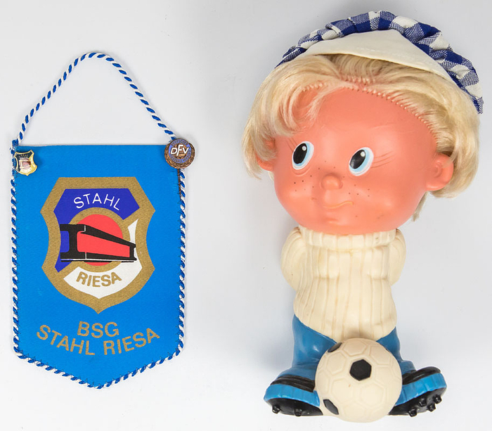 Maskottchen von Stahl Riesa aus den 1970er Jahren. Farbiger Kunststoff, mit Ku nsthaar und Stoffmütze. Höhe: 21 cm. Dabei: Kleiner Souvenirwimpel mit zwei Pins Stahl Riesa und Ehrennadel DFV. Alle aus den 1970er Jahren, 14x10 cm.