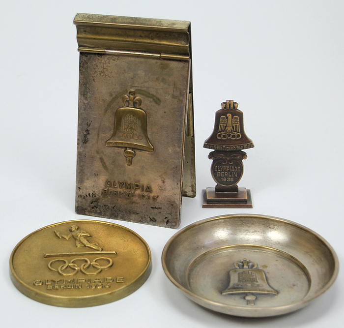  - Four different souvenirs from the 1936 Olympic Games: 1) Small silver-plated metal plate with the Olympic bell, 11.5 cm. 2) One-sided bronze plaque with relay runners and Olympic rings, 9.5 cm. 3) Small bronze sculpture of the 1936 Olympic bell with the inscription "Olympiade Berlin 1936", height: 8 cm. 4) Silver-plated metal clipboard with the Olympic bell and the embossed inscription "Olympia Berlin 1936", 15 x 8 cm Condition: A-B