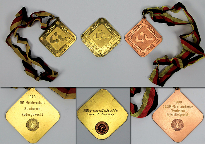  - Two winner's medals from the 1979 and 1980 East German Boxing Championships: 1) Gold medal inscribed "1979 East German Championship Senior Featherweight" with silk ribbon. 2) Bronze medal "1980 East German Championship Senior Light Middleweight" with silk ribbon. Each 8x8 cm in original box. Also included: an honorary plaque of the German Boxing Association (DBV) - East Germany with the inscription "Honorary Plaque Gerd Lang" on the reverse. Gilt bronze with an applied, enameled state coat of arms, 8.5x8.5 cm. --signs of usage Condition: B